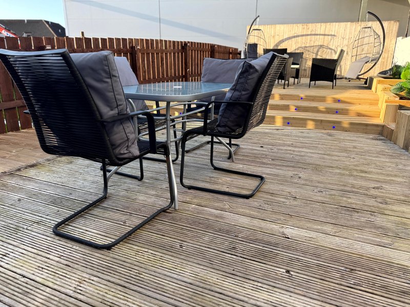 Outdoor patio with wooden decking, dining table, black chairs, and fenced garden area