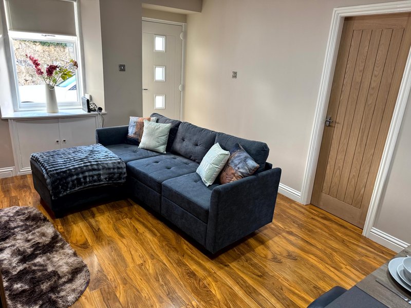 Modern living room with black corner sofa, wooden flooring, window with flowers, and wooden door