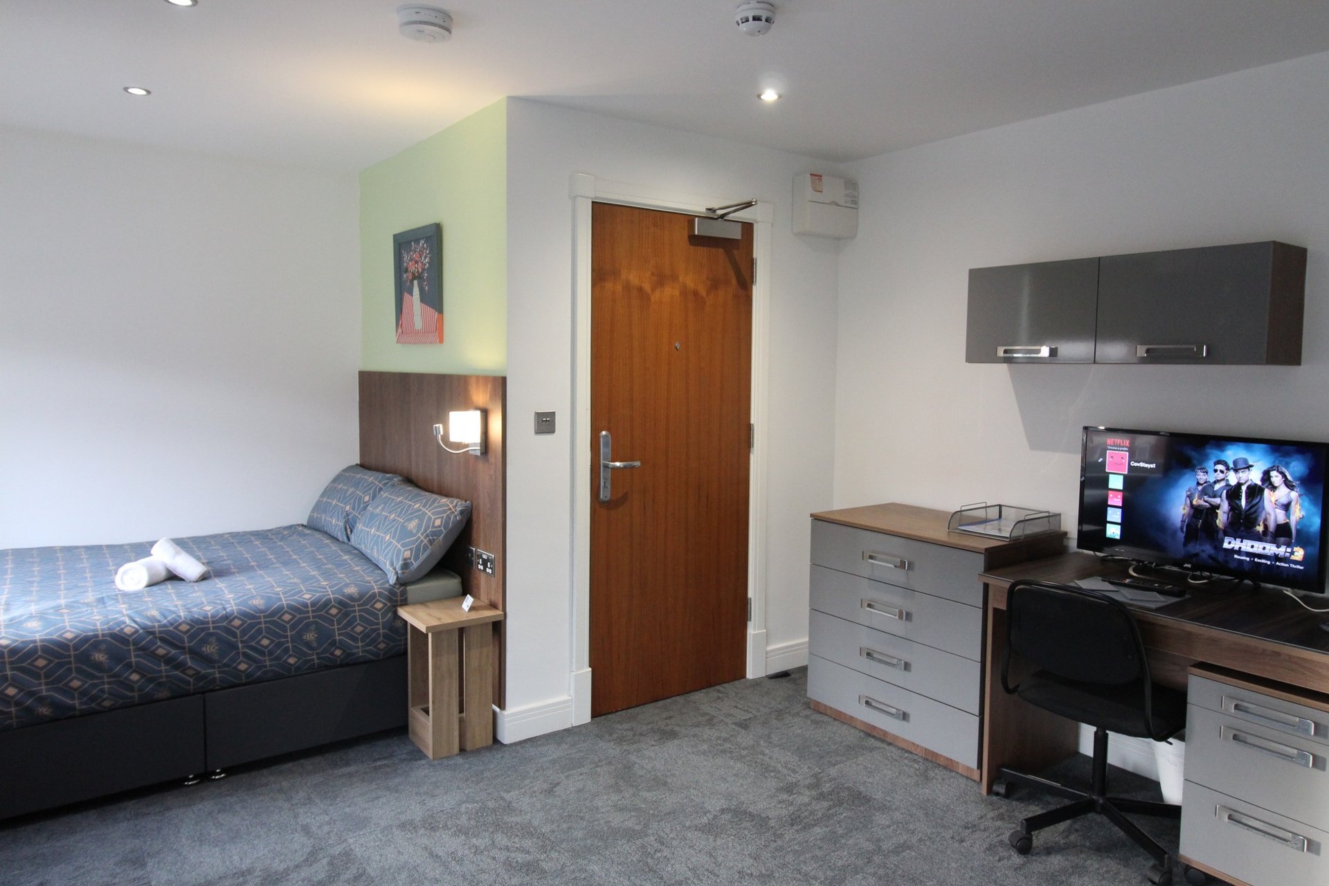 Bedroom with single bed, desk with monitor, storage drawers and wooden door