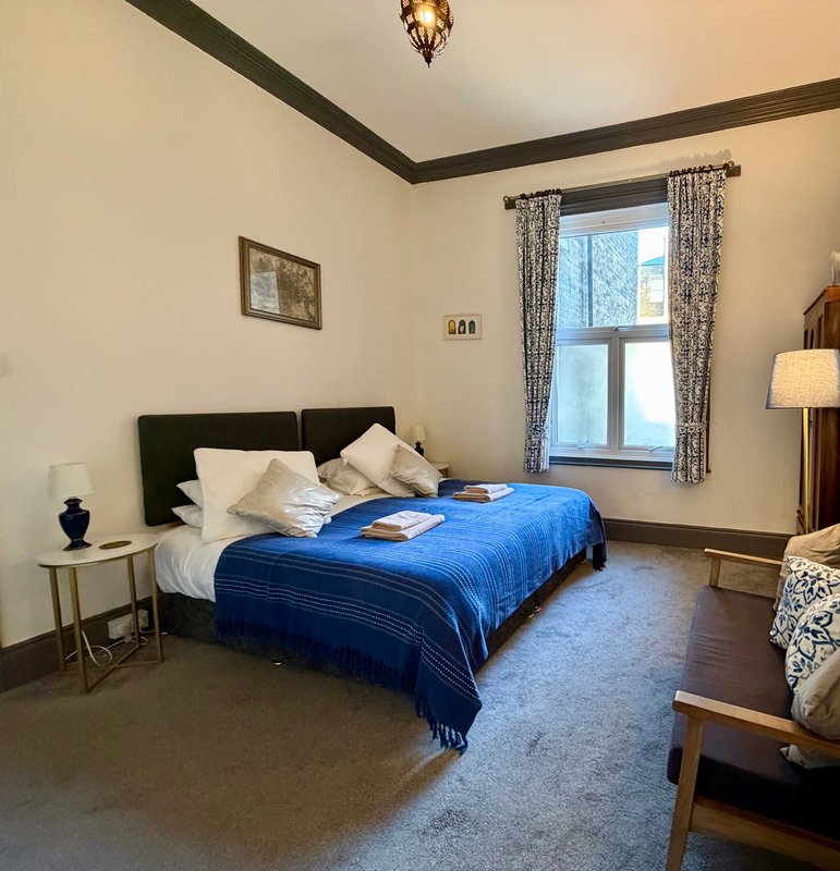 Bright bedroom with double bed, blue bedcover, black headboard, and large window with curtains