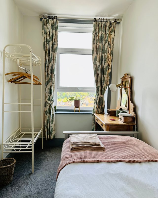 Compact bedroom with single bed, metal shelving unit, wooden desk, and patterned curtains