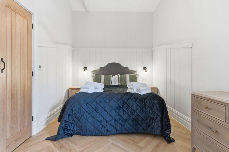 Bright bedroom with double bed, dark blue quilt, wooden headboard and white panelled walls