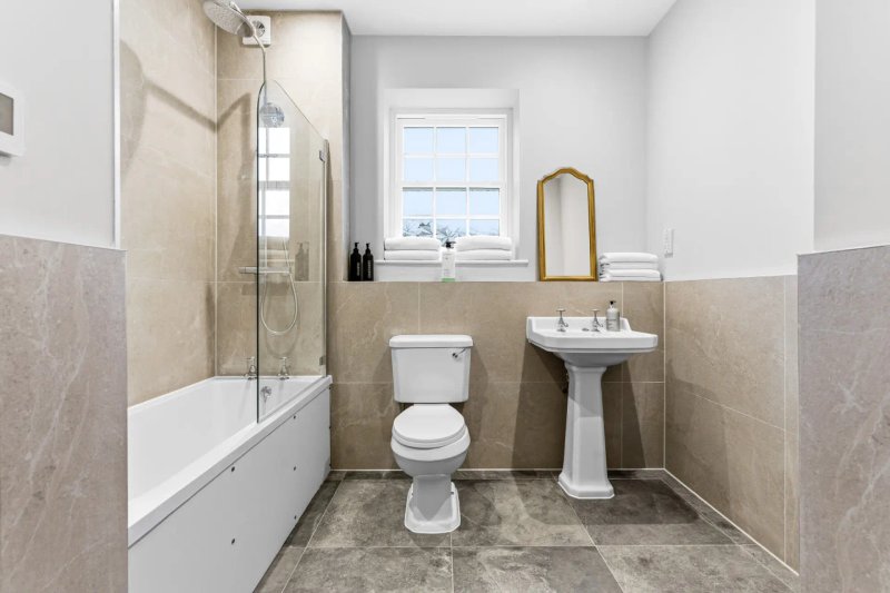 Clean bathroom with bathtub, pedestal sink, toilet, window and beige tile walls