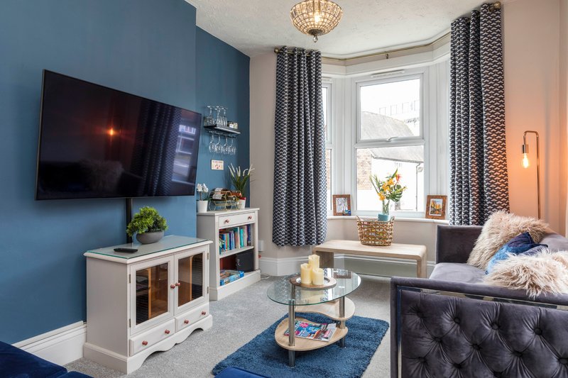 Bright living room with blue accent wall, TV, white storage unit, bay window with curtains and seating