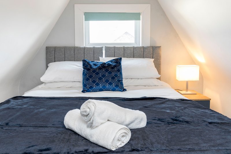 Attic bedroom with grey upholstered bed, rolled white towels, blue cushion and bedside lamp