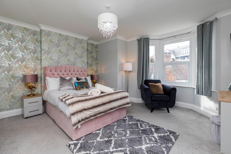 Modern bedroom with pink upholstered bed, patterned wallpaper, crystal chandelier and armchair by window