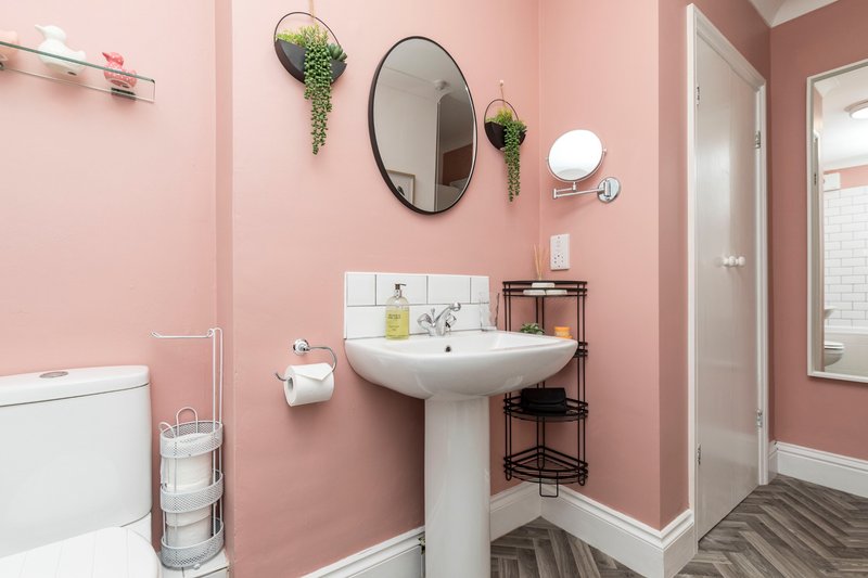 Pink bathroom with pedestal sink, oval mirror, hanging plants, toilet and shower enclosure