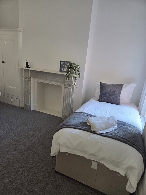 Bedroom with single bed, white pillows, gray carpet, fireplace mantle and wardrobe
