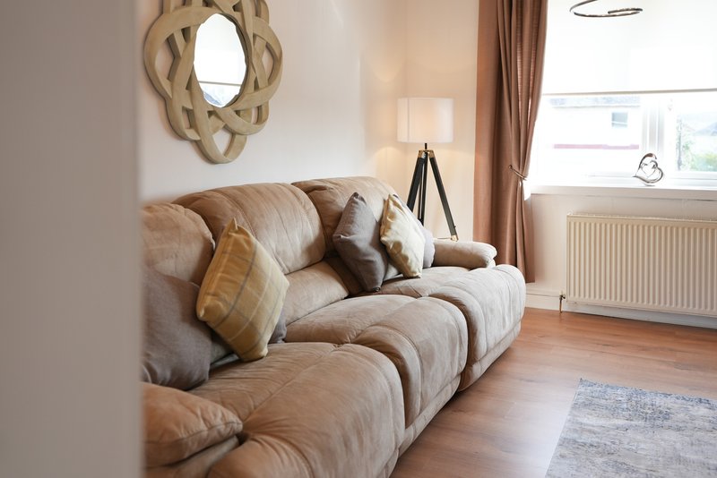 Living room with beige upholstered sofa, decorative cushions, wooden mirror, and natural light from windows