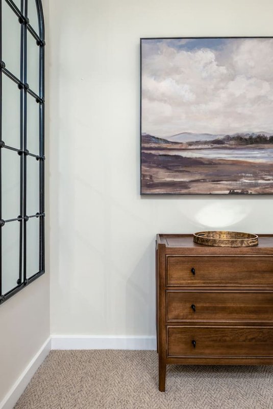 Bright corner bedroom with wooden chest of drawers, framed landscape painting, and gridded window