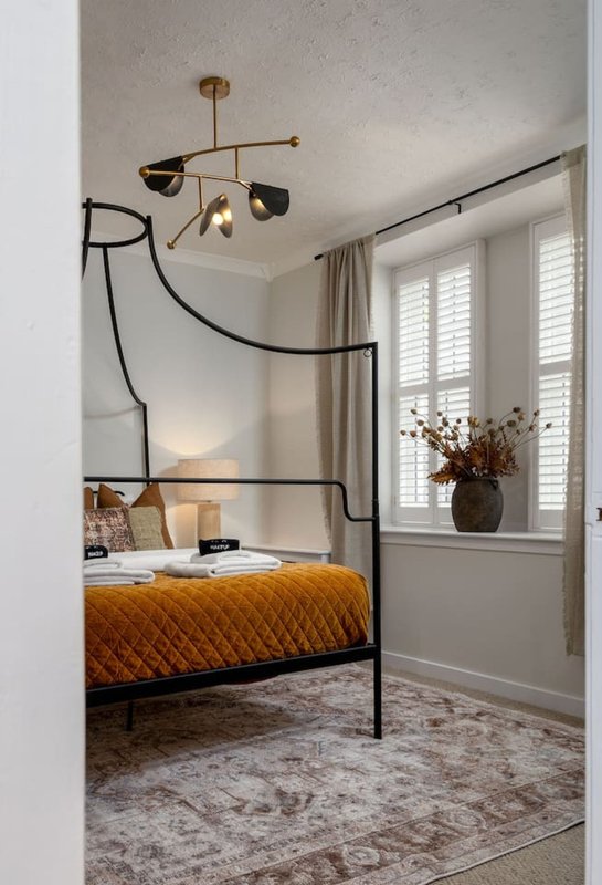 Bedroom with metal four-poster bed, mustard-colored bedding, brass pendant light, and shuttered windows