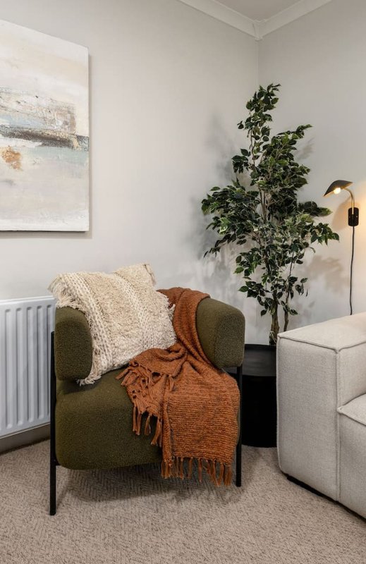 Modern living room corner with olive armchair, cream and terracotta throw blankets, potted plant, wall art, and floor lamp.