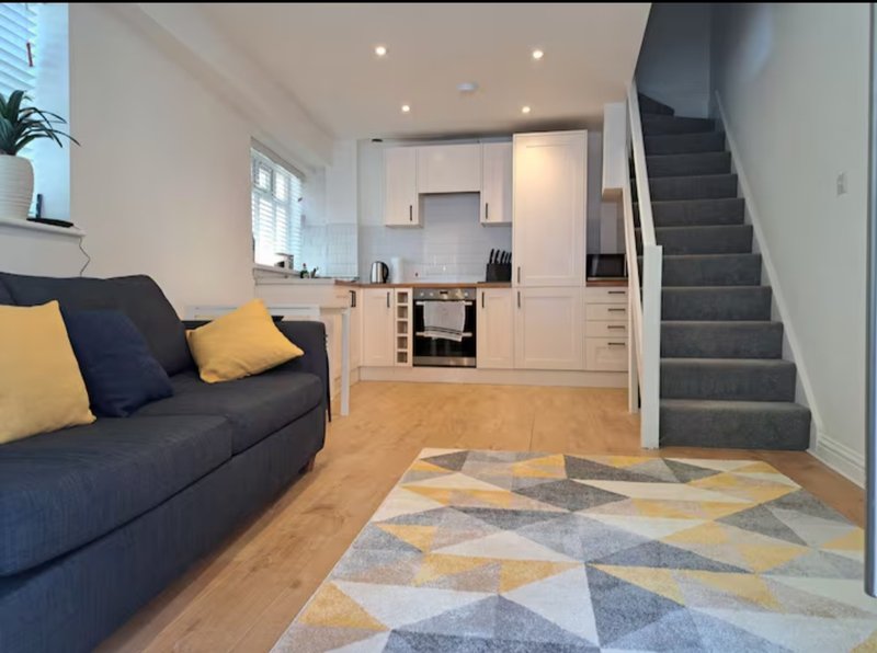 Open-plan living area with dark sofa, kitchen, stairs, geometric rug and wooden flooring