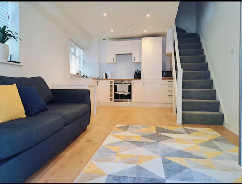 Open-plan living area with navy sofa, geometric rug, kitchen, and staircase to upper floor