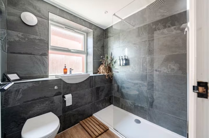 Modern bathroom with dark tile walls, white toilet, vanity sink, walk-in shower and wooden floor accent
