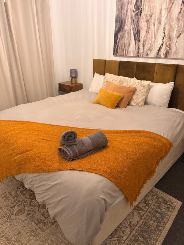 Bedroom with king bed, orange blanket, cushions, wooden headboard and bedside lamp