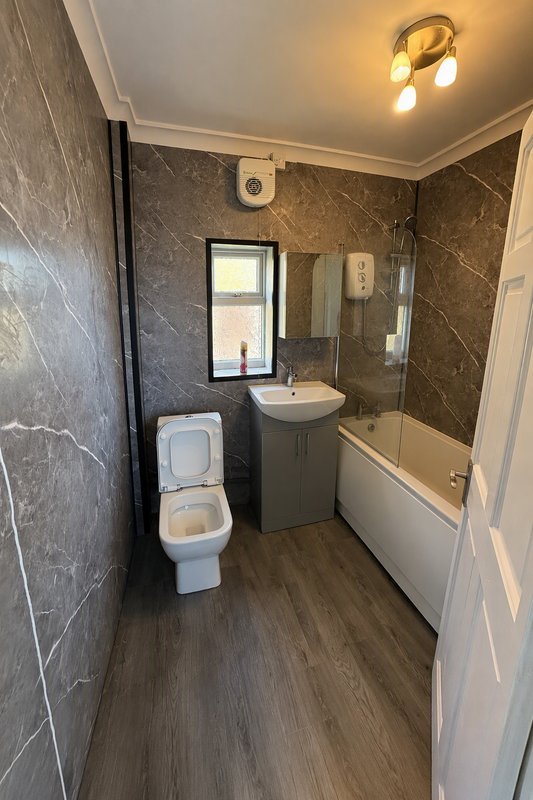 Modern bathroom with dark tile walls, white toilet, sink vanity, bathtub with glass screen, and window
