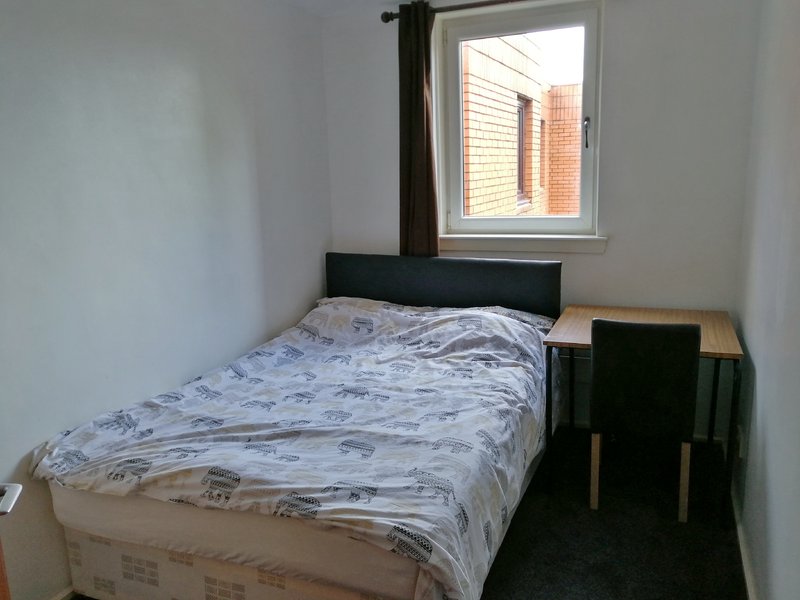 Bedroom with single bed, black headboard, wooden chair, and window overlooking brick building
