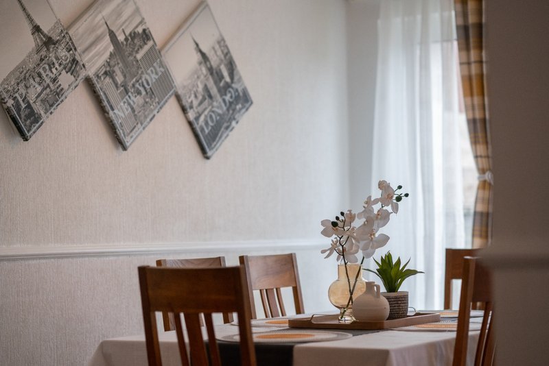 Dining area with wooden chairs, table, vase with dried flowers, and framed architectural prints on wall