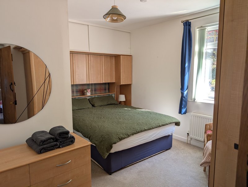 Bedroom with double bed, green bedding, wooden headboard storage, round mirror and dresser