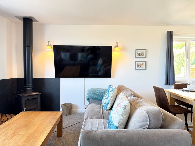 Open-plan living room with wood-burning stove, grey sofa, wall-mounted TV, and dining area