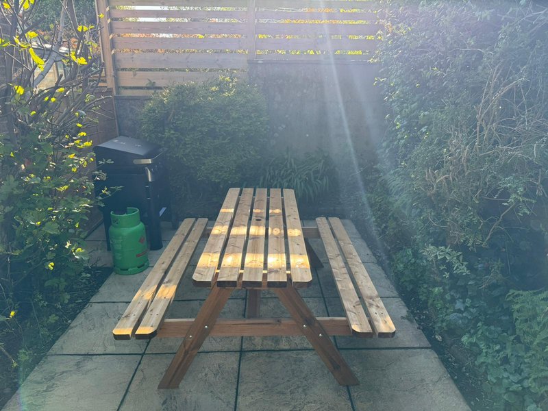 Wooden picnic table in garden with green bin and overgrown vegetation