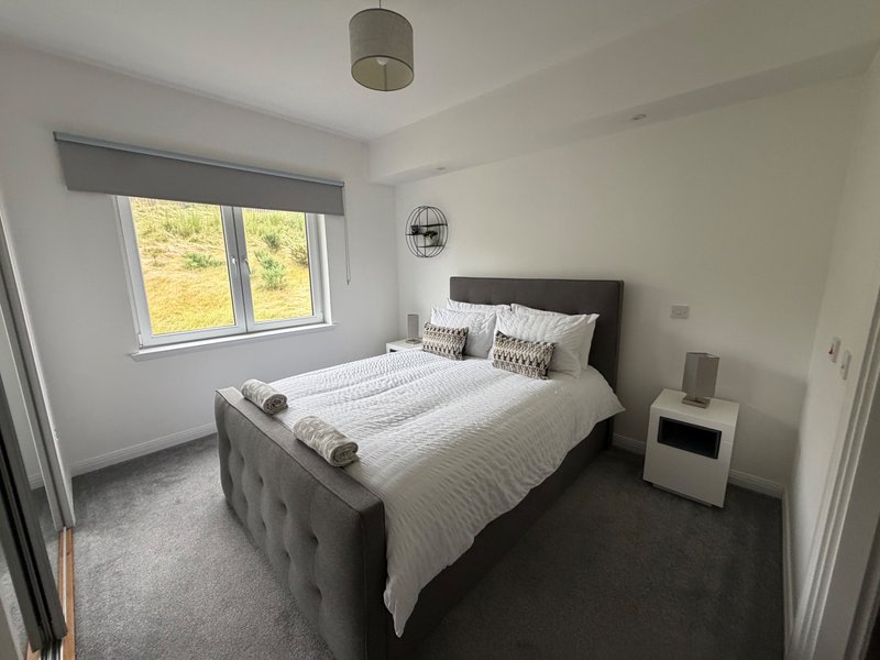 Bright modern bedroom with double bed, grey headboard, white bedding, and window with countryside view