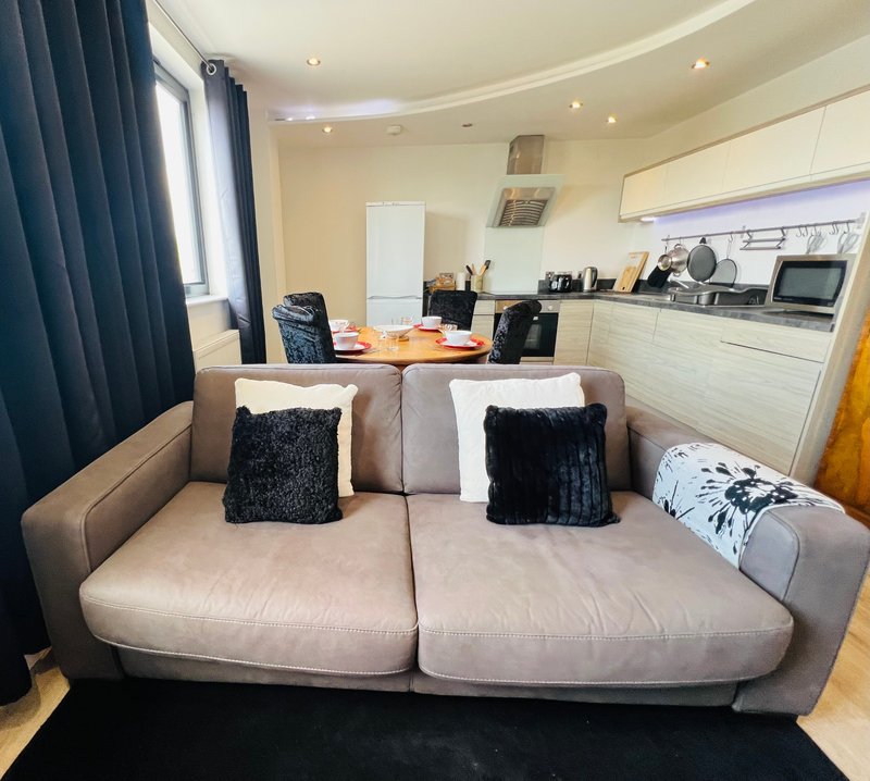 Open-plan living area with beige sofa, cushions, kitchen units and window with blue curtains