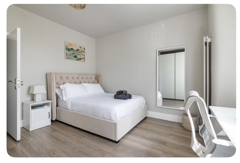 Bright modern bedroom with beige upholstered bed, white bedding, wooden flooring and mirrored wardrobe