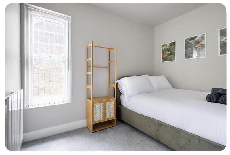 Bright bedroom with double bed, wooden shelving unit, window with radiator, and framed landscape photos