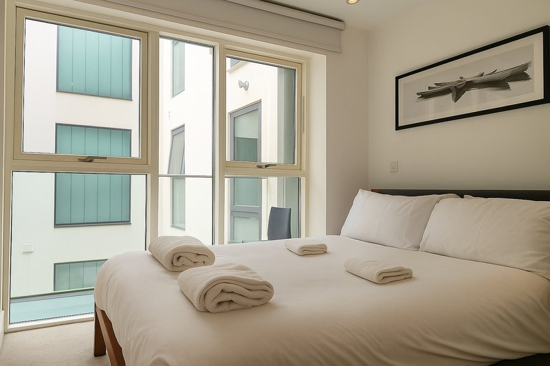 Bright modern bedroom with double bed, folded towels, and floor-to-ceiling windows overlooking building