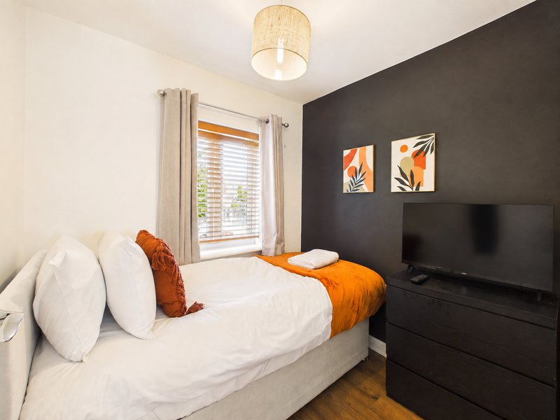 Modern bedroom with double bed, white bedding, orange throw, dark accent wall and TV