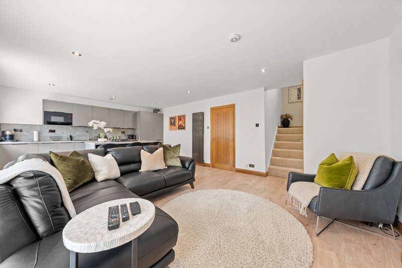 Open-plan living room with black leather sofa, accent chairs, cream rug and kitchen area