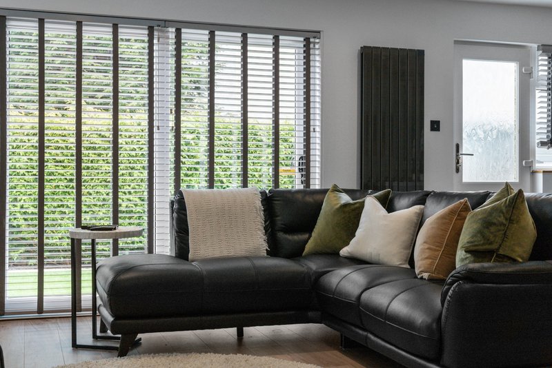 Modern living room with black leather sectional sofa, decorative cushions, and floor-to-ceiling shuttered doors overlooking garden