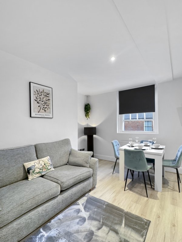 Modern living room with gray sofa, small dining table, framed artwork, and window with blind