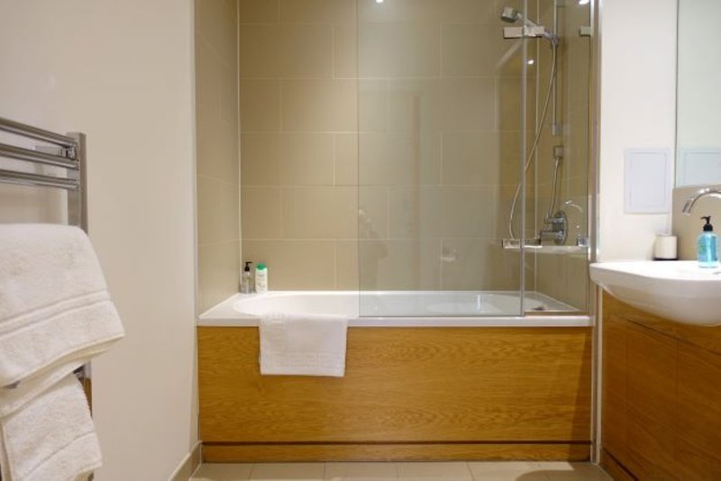 Bathroom with wooden bath panel, glass shower screen, beige tiles and white towels