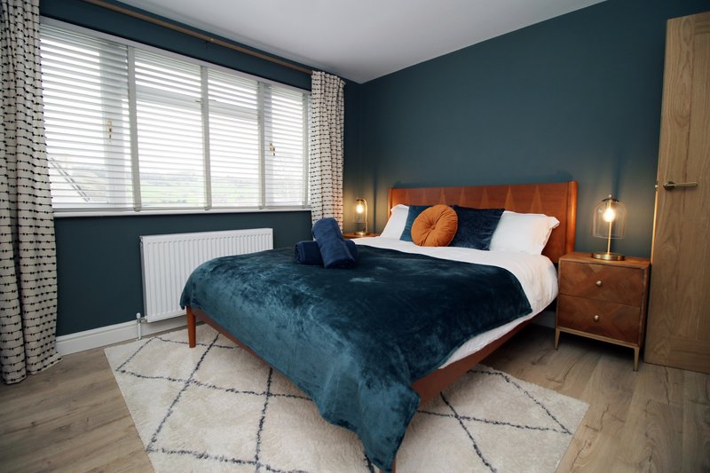 Modern bedroom with double bed, orange headboard, teal accent wall, wooden nightstand, and large window with shutters
