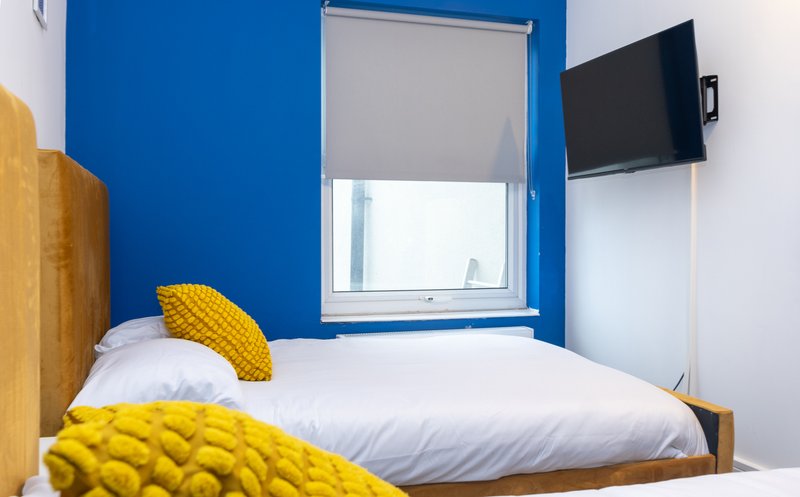 Compact bedroom with single bed, blue walls, yellow cushions and wall-mounted TV