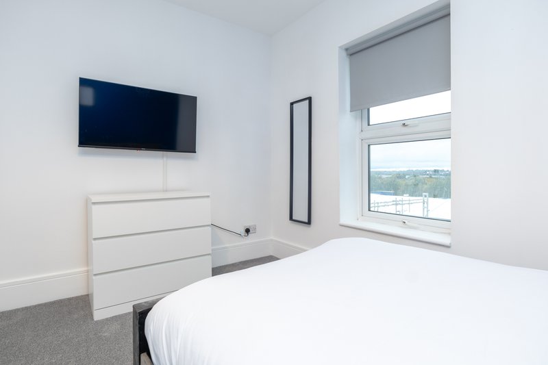 Modern bedroom with white bed, wall-mounted TV, dresser and window overlooking green landscape