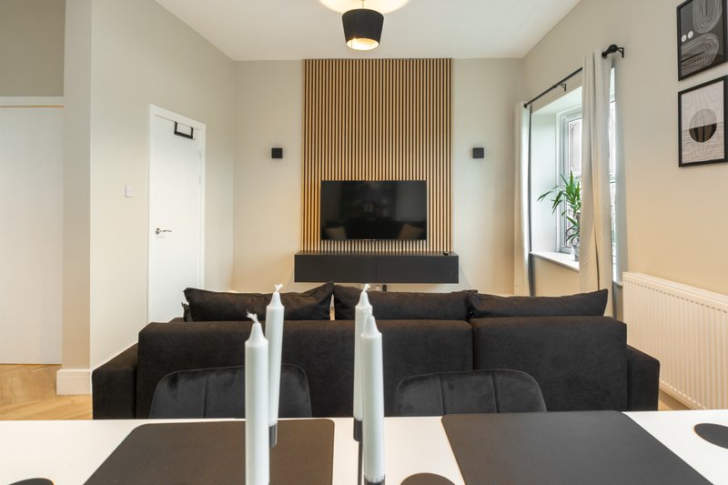 Modern living room with black sofa, mounted TV on wooden feature wall, and dining table in foreground