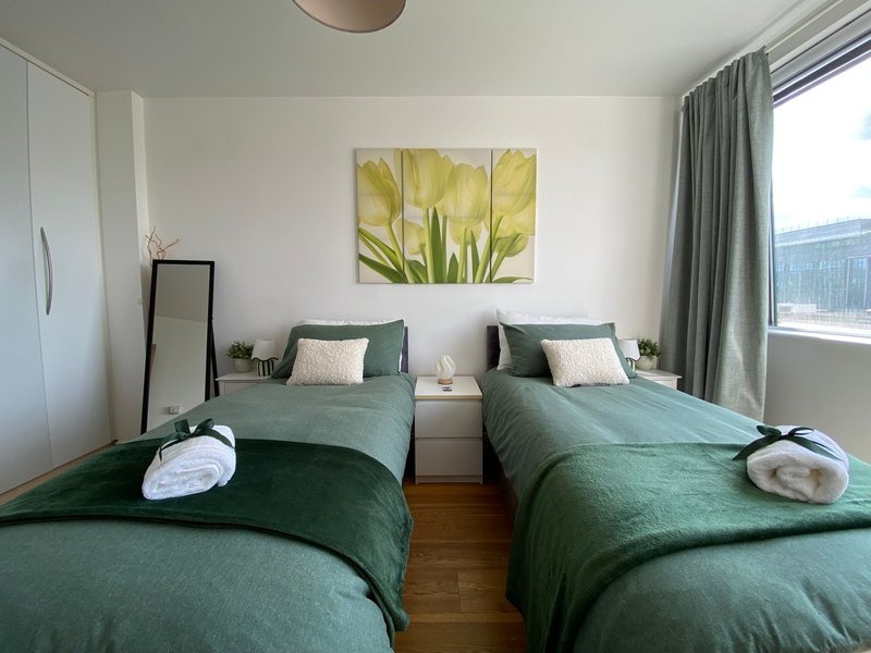 Bright twin bedroom with green bedding, white pillows, floral artwork, and large windows with grey curtains