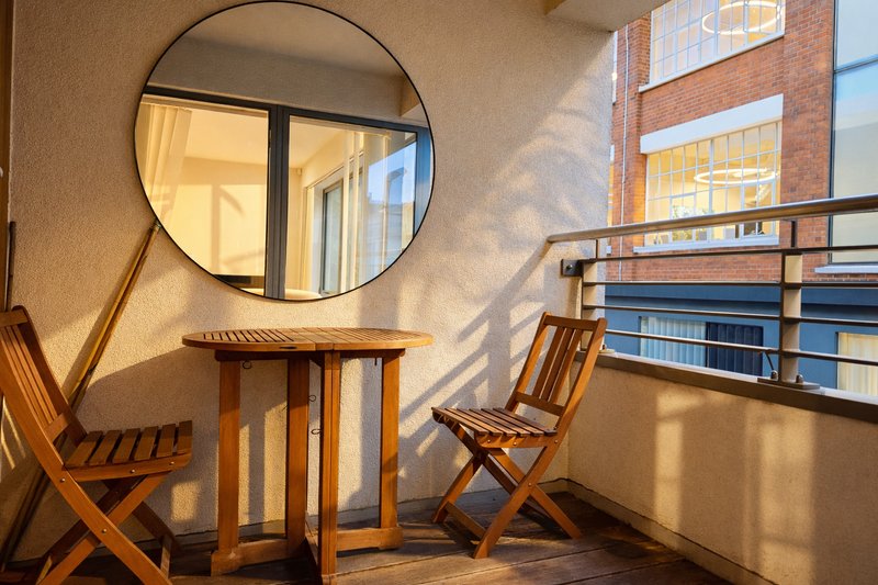 Bright balcony with wooden chairs, small table, and round mirror overlooking urban buildings