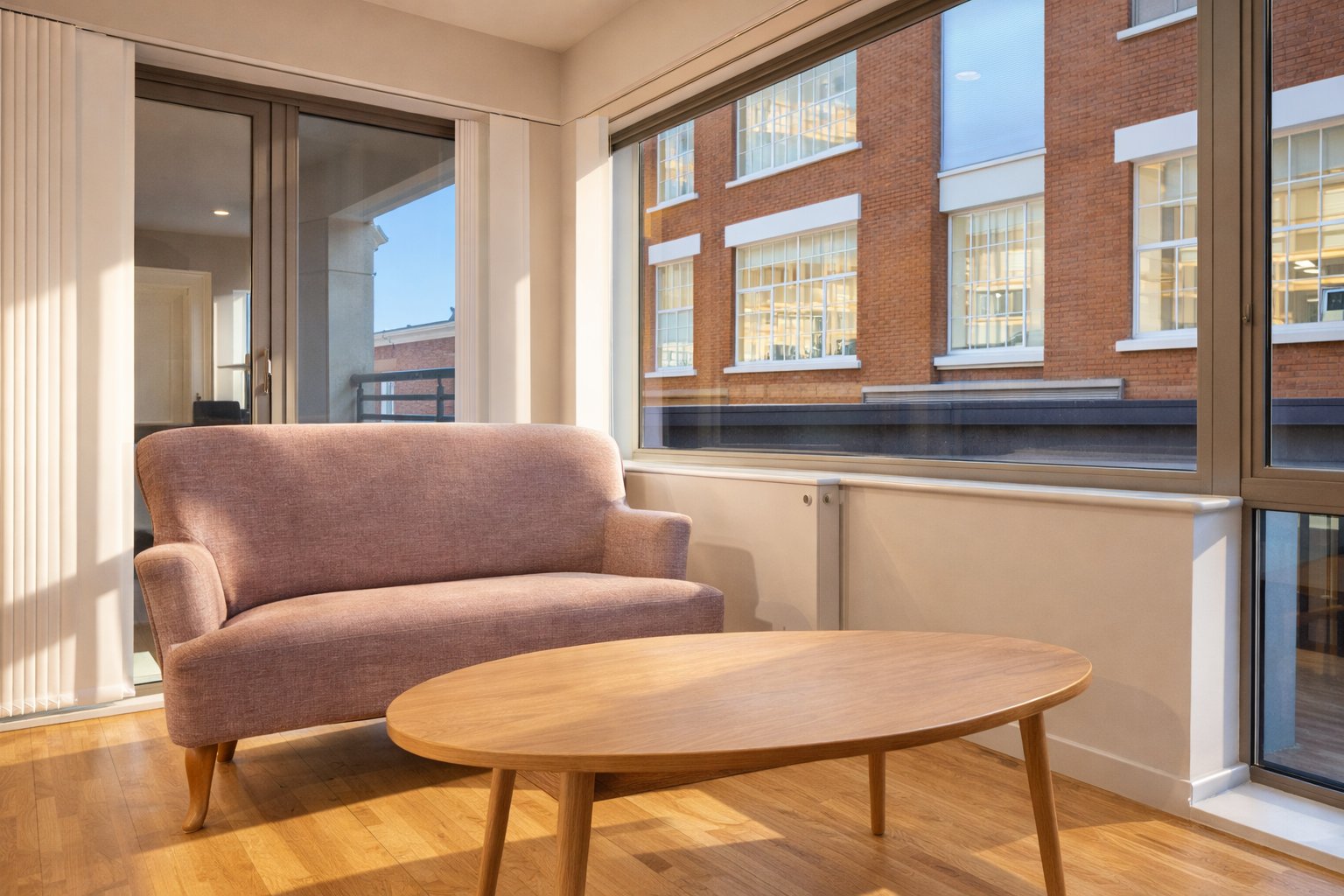 Bright living room with mauve sofa, wooden coffee table, large windows overlooking brick buildings