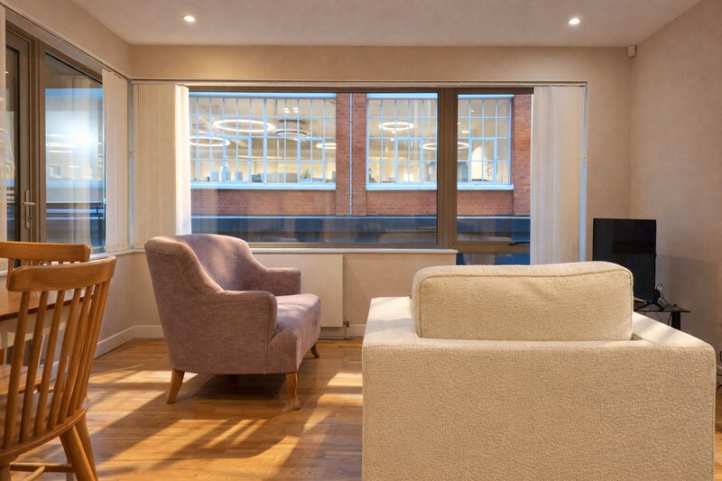 Bright living room with upholstered sofa, armchair, wooden chair and large windows overlooking brick building
