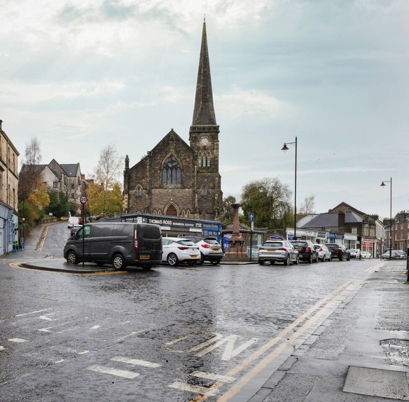 Town square with parked cars, stone church with spire, wet pavement and street lamps