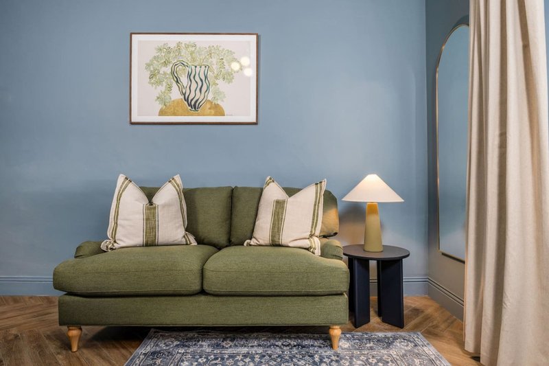Living room with green upholstered sofa, decorative pillows, side table with lamp and framed botanical artwork