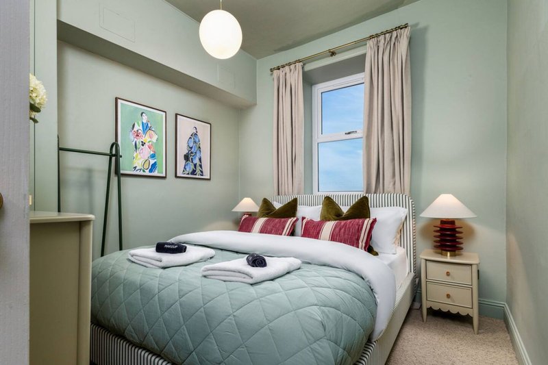 Bedroom with double bed, sage green walls, framed artwork, window with curtains and bedside lamp