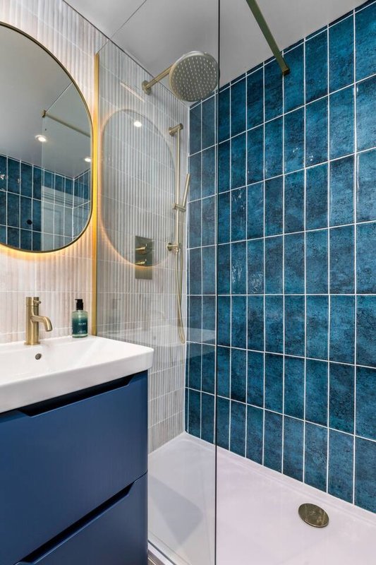 Modern bathroom with blue tile wall, white sink, gold-rimmed mirror and glass shower enclosure