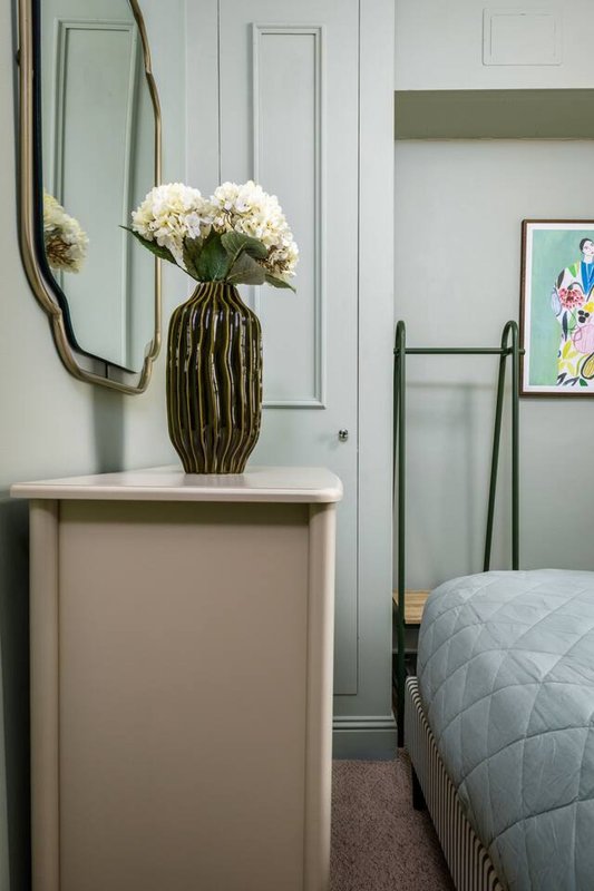 Bedroom corner with white dresser, striped vase with white flowers, mirror, and bed with blue bedding