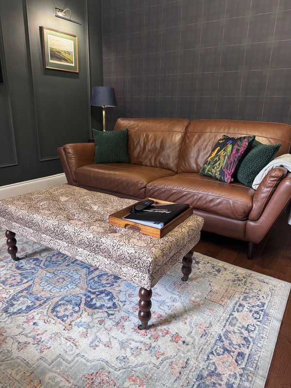 Living room with brown leather sofa, patterned ottoman, blue and cream rug, and framed artwork on wall